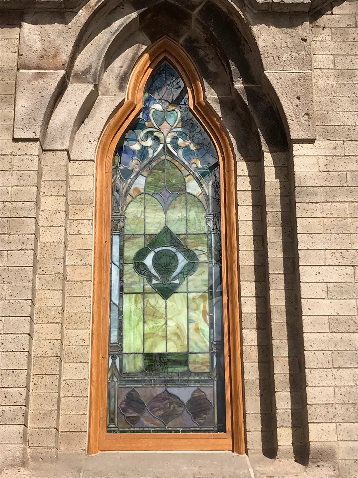 Arched stained glass window with blue and green panels in wooden frame set in stone wall