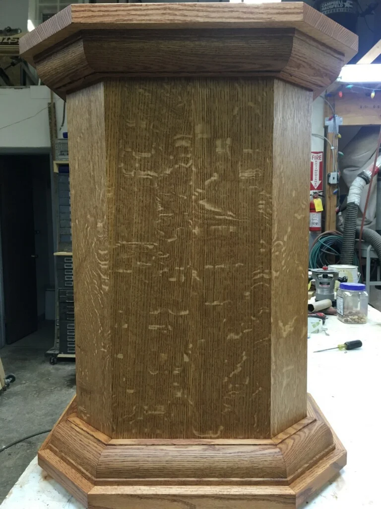 Wooden pedestal with carved relief design, crown molding top, and beveled base in workshop